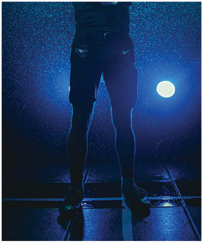 A person wearing a football uniform with the number nine and a headband is holding a football and pointing forward while standing in a dramatic, rain-like setting with bright lights behind them. Description generated by AI