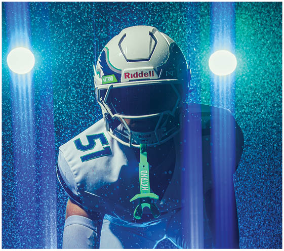 A person wearing a football uniform and helmet is crouched in a ready stance under blue lighting with rain or mist falling around them. Description generated by AI