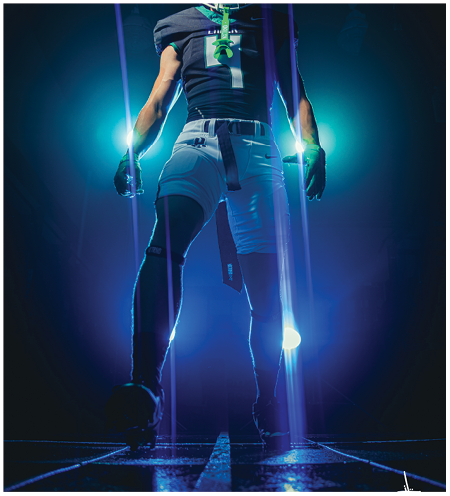 A person wearing a football uniform with a helmet and gloves stands confidently with glowing blue lights behind them and purple light streaks in the foreground. Description generated by AI