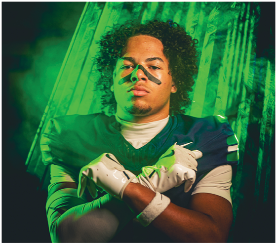 A person with curly hair wearing a sports jersey and gloves poses with arms crossed and black athletic tape under their eyes against a green-lit background. Description generated by AI