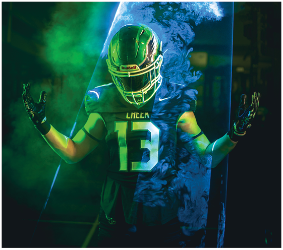 A person wearing a football helmet and jersey with the number 13 stands with arms raised amid swirling green and blue smoke and dramatic lighting. Description generated by AI