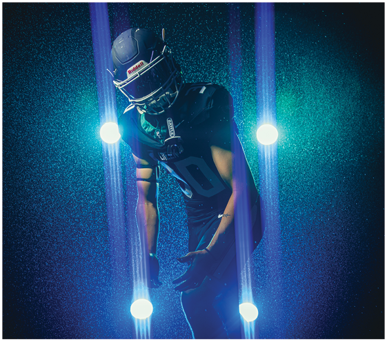A person wearing a dark football uniform and helmet is standing with arms bent in front of a background illuminated by four vertical beams of blue light. Description generated by AI