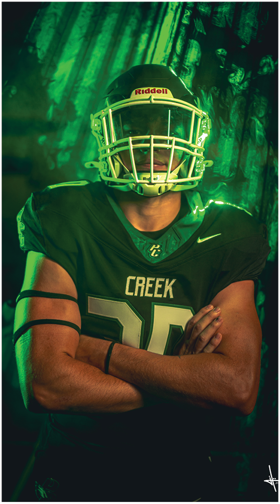 A person wearing a dark green football uniform with the word Creek on the chest and a helmet stands with arms crossed against a green-lit background. Description generated by AI