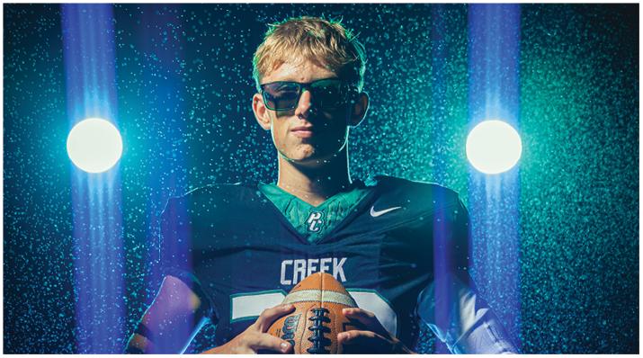 A person wearing sunglasses and a football jersey with the word “CREEK" holds a football under dramatic lighting with water droplets in the air. Description generated by AI