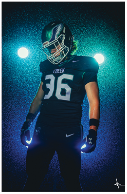 A person wearing a dark football uniform with the number 36 and the word Creek on the jersey stands with head slightly bowed against a dramatic blue-lit background with two bright lights behind. Description generated by AI