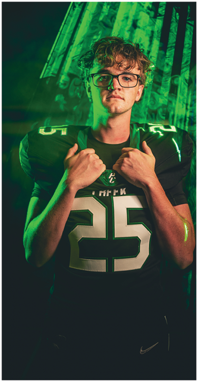 A person wearing glasses and a dark football jersey with the number 25 stands confidently with hands gripping the collar against a dark background with green lighting. Description generated by AI