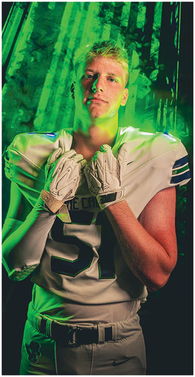 A person wearing glasses and a dark football jersey with the number 25 stands confidently with hands gripping the collar against a dark background with green lighting. Description generated by AI