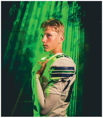 A person with short hair wearing a white sports jersey with dark stripes poses thoughtfully against a green-lit background. Description generated by AI