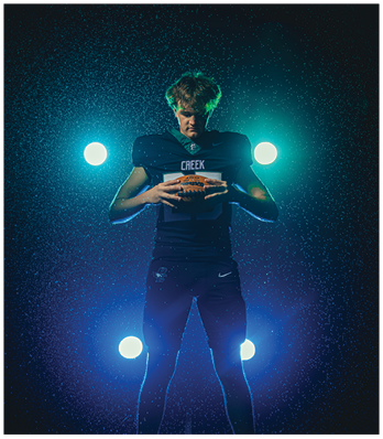A person wearing a dark football uniform with the word Creek on the chest is standing in a dramatic rain-lit scene holding a football with bright lights behind them. Description generated by AI