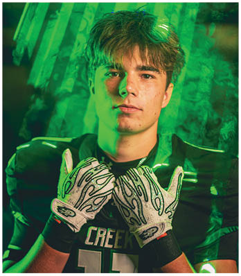 A person wearing a football helmet and uniform with crossed arms stands in front of a textured background illuminated by green and blue lighting. Description generated by AI