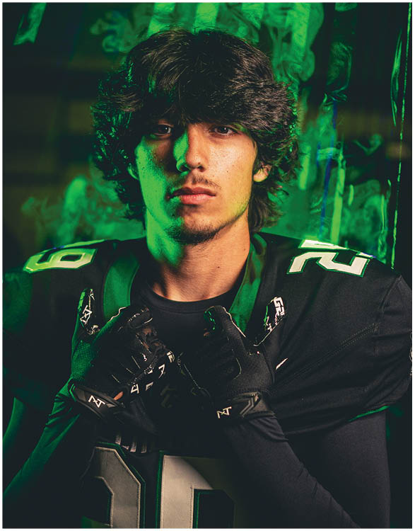 A person with medium-length wavy hair wearing a black sports jersey and gloves poses confidently against a dark background with green lighting. Description generated by AI