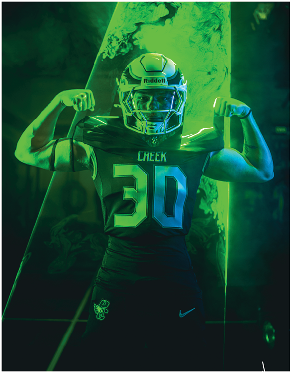 A person wearing a football helmet and jersey with the number 30 flexes their arm muscles in front of a green-lit background. Description generated by AI
