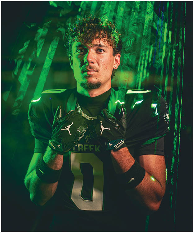 A person wearing a football uniform with the number 11 holds a football helmet in front of their face while standing against a dark, smoky background with blue and green lighting. Description generated by AI
