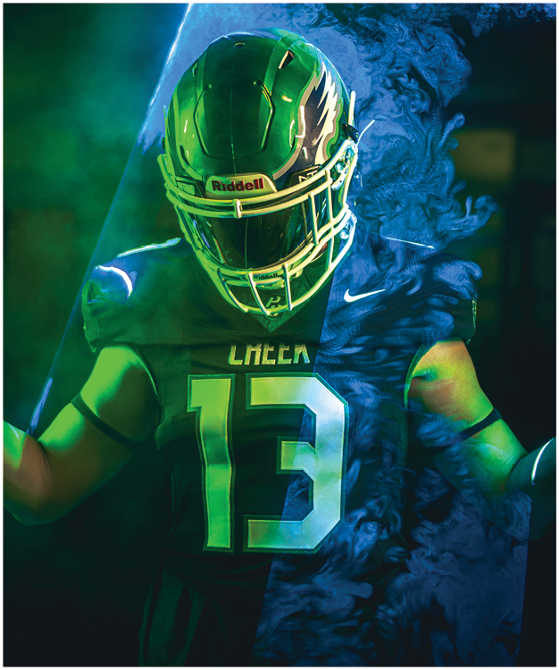 Person with shoulder-length hair wearing a football jersey with the number 55 stands with hands clasped near the chest against a dark background with green lighting effects. Description generated by AI