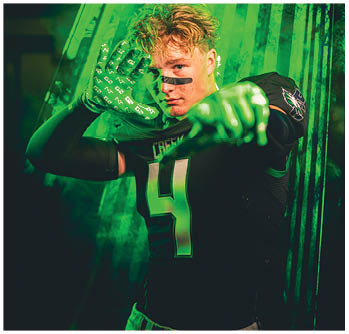 A person with curly hair wearing a sports jersey and gloves poses with one hand raised and the other pointing forward against a green smoky background. Description generated by AI