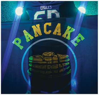 A person wearing a sports jersey with the number 50 and the word Eagles stands behind a flag that says Pancake with an image of stacked pancakes, illuminated by bright lights and blue light beams. Description generated by AI