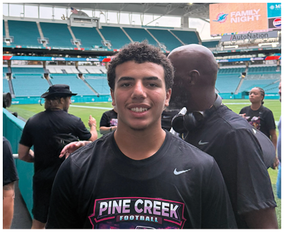 A person wearing a Pine Creek Football shirt is smiling in a stadium with several other people in the background near the field. Description generated by AI