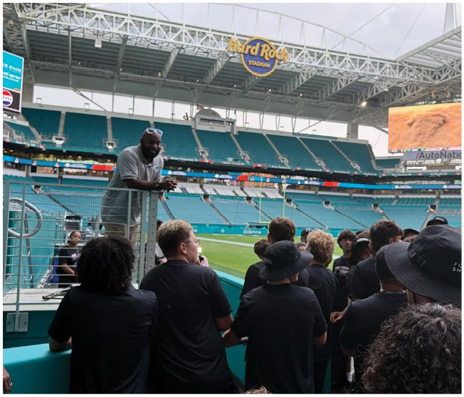 A group of people wearing black clothing are gathered inside a large stadium near the field, listening to a person standing on a platform speaking to them. Description generated by AI