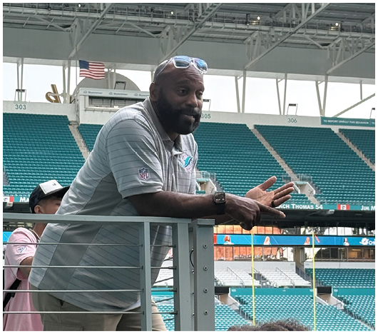 A person wearing a light gray polo shirt with a sports logo and sunglasses on their head is standing in front of empty stadium seats under a Hard Rock Stadium sign. Description generated by AI