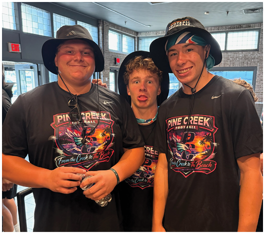 Three adults wearing matching Pine Creek Football t-shirts and hats pose together indoors with one making a playful face. Description generated by AI