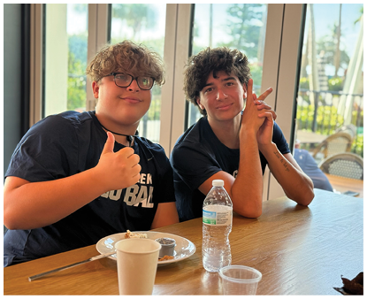 Two people with curly hair sit at a table with plates and cups, one giving a thumbs-up and the other resting their chin on their hand, in a bright room with large windows. Description generated by AI
