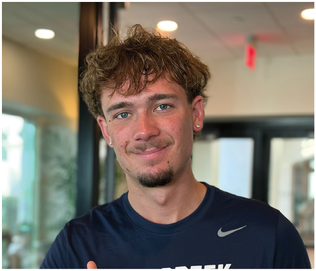 A person with curly hair and earrings is smiling softly while wearing a dark blue Nike shirt in an indoor setting with blurred background. Description generated by AI
