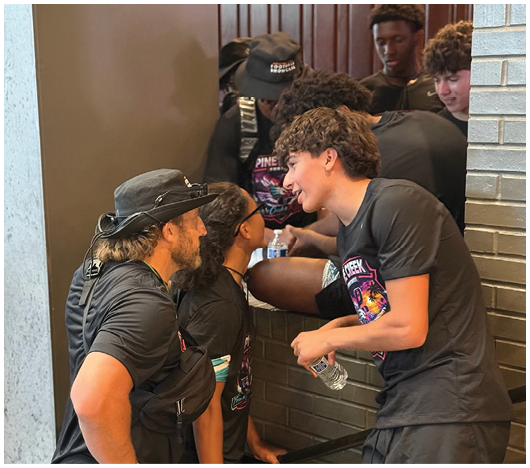A group of people wearing matching black shirts are gathered closely together, some leaning in and holding water bottles, engaged in conversation near a brick wall. Description generated by AI