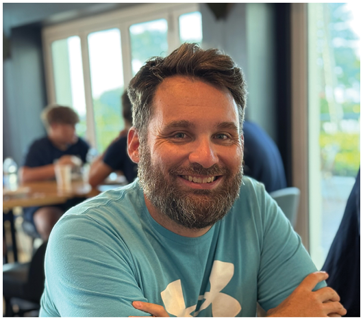 A person with a beard and short hair is smiling and sitting with arms crossed in a casual setting with other people and large windows in the background. Description generated by AI