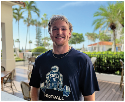 A person with short curly hair wearing a dark blue t-shirt with a football graphic and the word Eagles is smiling while standing outdoors on a patio with palm trees and chairs in the background. Description generated by AI