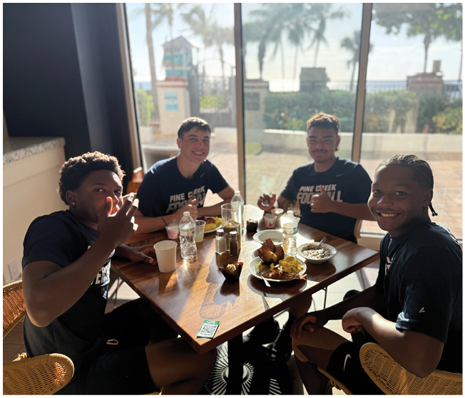 Four people wearing matching dark sports shirts are sitting around a wooden table with food and drinks, smiling and giving thumbs up in a sunlit room with large windows showing palm trees outside. Description generated by AI