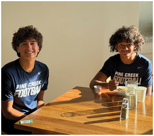 Two people with curly hair wearing matching Pine Creek Football shirts are sitting at a wooden table with food and drinks, smiling in a sunlit room. Description generated by AI