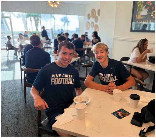 Two people wearing matching dark blue Pine Creek Football shirts sit at a table with drinks and snacks in a bright cafe filled with other people. Description generated by AI