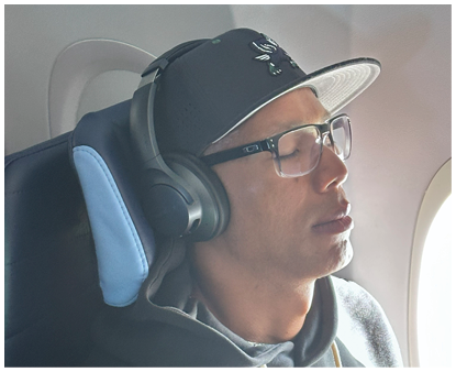 A person wearing glasses, a cap, headphones, and a hoodie with the text “Pine Creek Football" is sitting by an airplane window. Description generated by AI