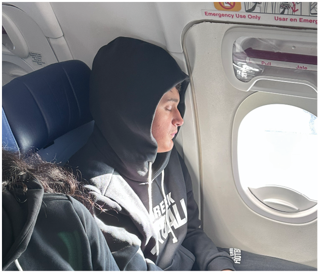 Two people wearing matching black hoodies and sweatpants with “Pine Creek Football" printed on them are sitting next to each other inside an airplane near a window. Description generated by AI