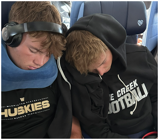 Two people wearing black sportswear with “Pine Creek Football" and "Huskies" logos are resting closely together, one with headphones and a neck pillow. Description generated by AI
