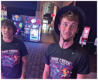 Two people wearing matching Pine Creek Football t-shirts stand indoors near arcade machines, with one person wearing a wide-brimmed hat and smiling. Description generated by AI