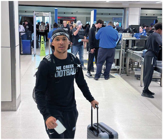 A person wearing a headband and a black shirt with “CREEK FOOTBALL" printed on it stands in the foreground of an airport security checkpoint with several people in line and security staff in the background. Description generated by AI
