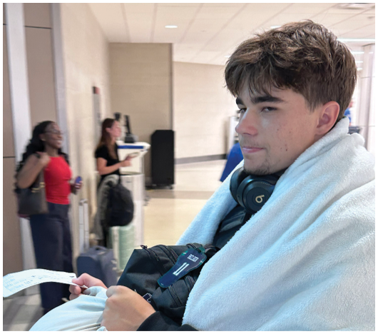A person wrapped in a white blanket is sitting with headphones around their neck, holding a pillow and a piece of paper, while two other people stand in the background near luggage. Description generated by AI