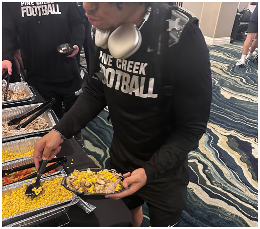 A person wearing a black long-sleeve shirt with “Pine Creek Football" printed on it is serving food from a buffet tray onto a plate. Description generated by AI