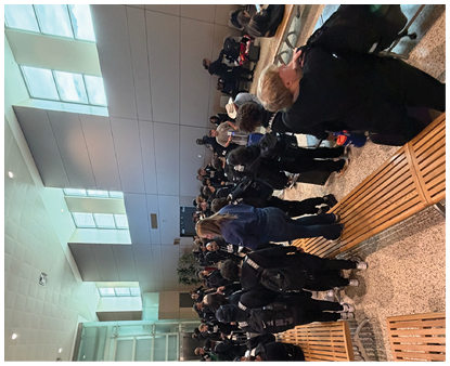 A group of people dressed mostly in dark clothing are gathered indoors near wooden benches and a large window with natural light. Description generated by AI