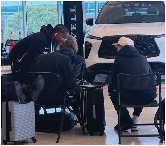 Two people sitting on chairs in front of a white vehicle with the word “JEWELL" visible on the windshield inside a modern building. Description generated by AI