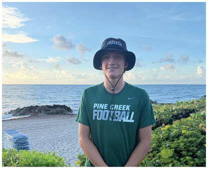 A person wearing a green Pine Creek Football shirt and a black bucket hat is smiling outdoors near a body of water with greenery in the background. Description generated by AI
