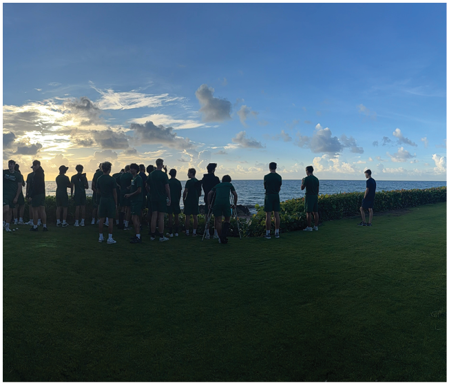A group of people wearing dark clothing stand on a grassy area near the ocean at sunset with a partly cloudy sky. Description generated by AI