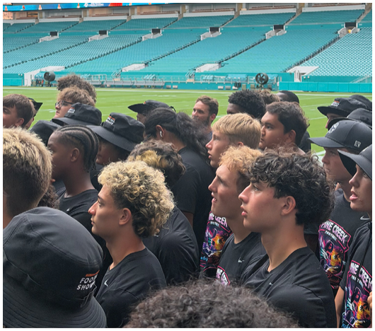 A group of people with varied hairstyles and hats are gathered closely together inside a large empty stadium with teal seats and a green field. Description generated by AI
