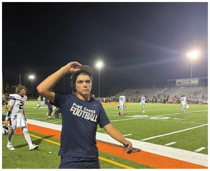 A person wearing a dark blue shirt and cap stands on the sidelines of a lit football field at night while other people in football uniforms are on the field and near the stands. Description generated by AI