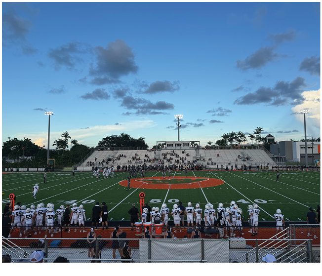 A football game is taking place on a field with players in white uniforms on the sidelines and a crowd seated in the bleachers under a partly cloudy sky at dusk. Description generated by AI