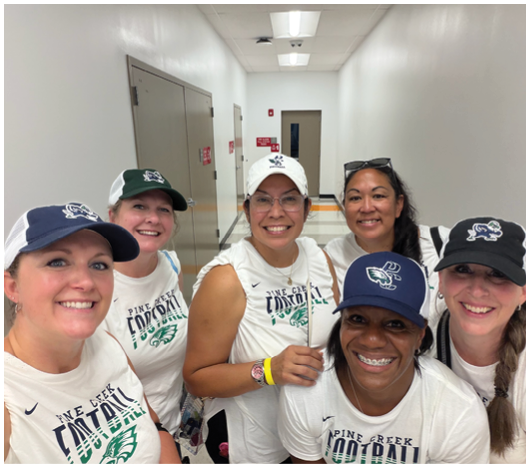 A group of six smiling adults wearing matching white sleeveless shirts and baseball caps pose together in a hallway. Description generated by AI