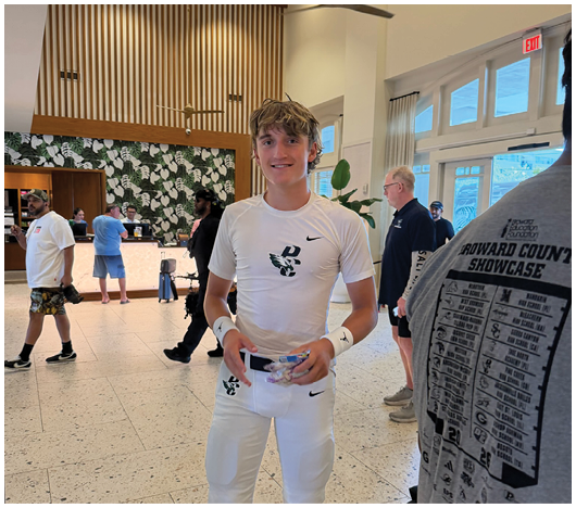 A person with tousled hair wearing a white Nike shirt stands smiling in a modern indoor space with several other people in the background near a counter and tropical-themed wall decor. Description generated by AI
