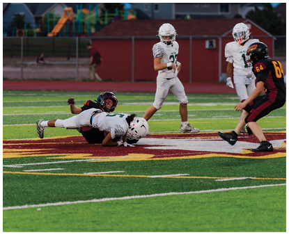Two people in football uniforms are on the ground in the middle of a field while three others in football uniforms stand nearby during a game. Description generated by AI