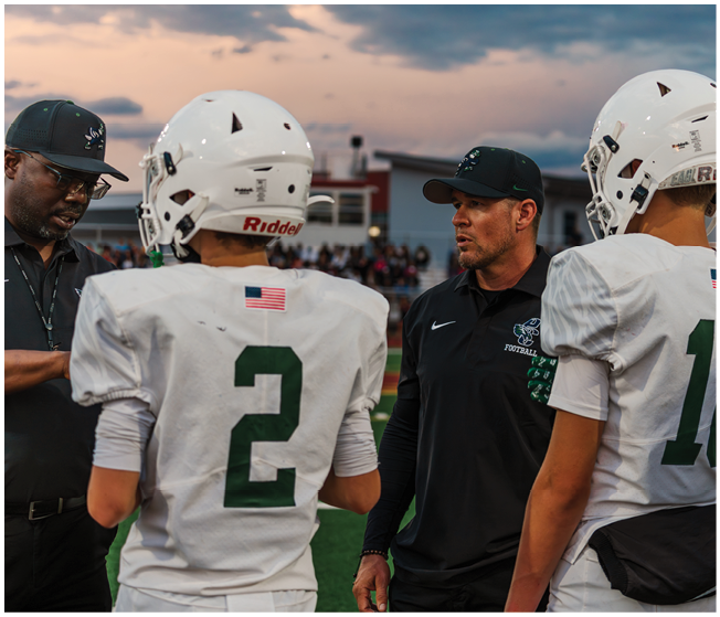Two adults in black sportswear and two people in white football uniforms with helmets are standing on a field during sunset, seemingly engaged in conversation. Description generated by AI